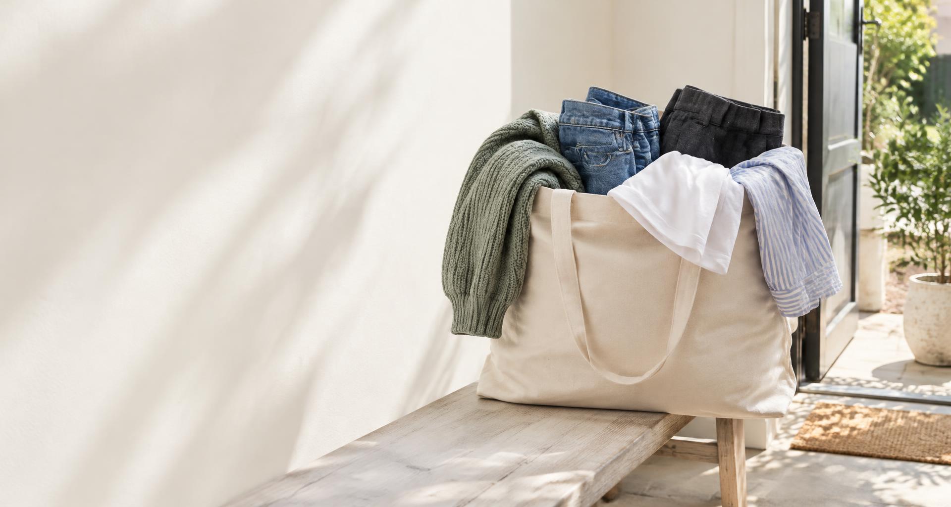 A canvas bag filled only with everyday clothes: a sage jumper, jeans, charcoal work trousers, a white T-shirt and a striped shirt